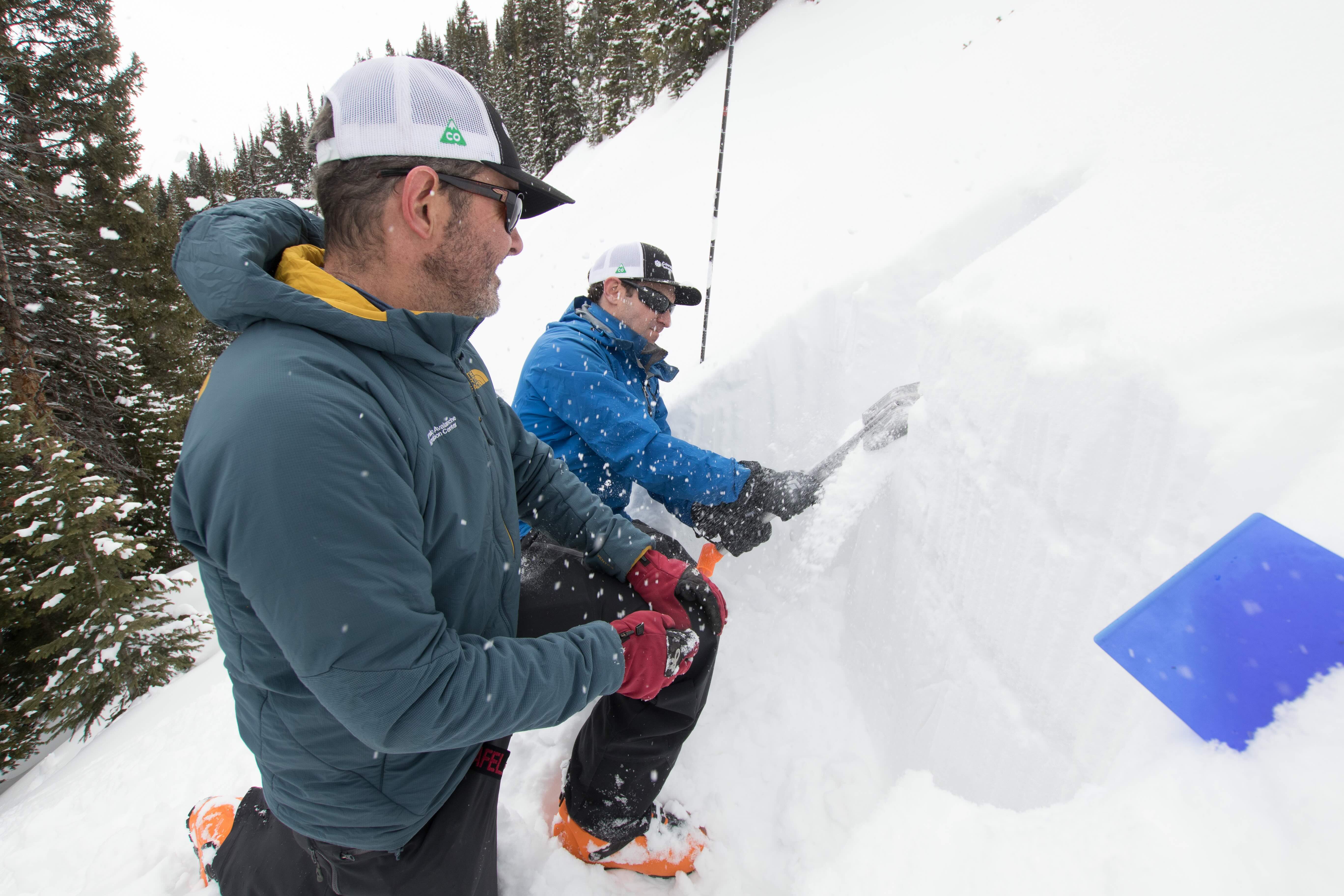 Two people wearing winter gear kneel in the snow while examining a snow pit during an avalanche safety field exercise. One uses a shovel to assess snow layers as part of a backcountry snowpack evaluation.