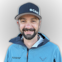 John Rizza, CAIC Field Technician Central Mountains at CAIC, smiling and wearing a blue jacket against a light grey background.